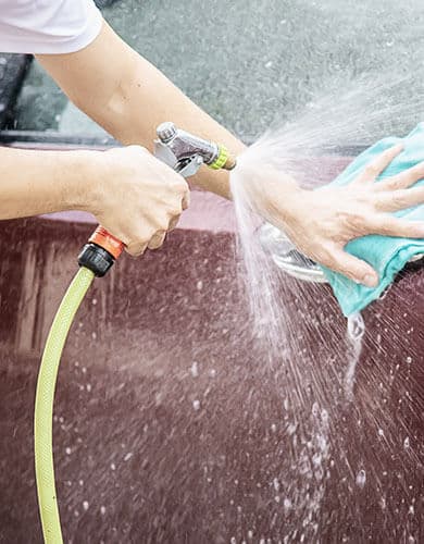 Picture of a car being rinsed with a water hose and scrubbed with a blue microfiber towel. This is similar to how we perform our mobile car washing services.